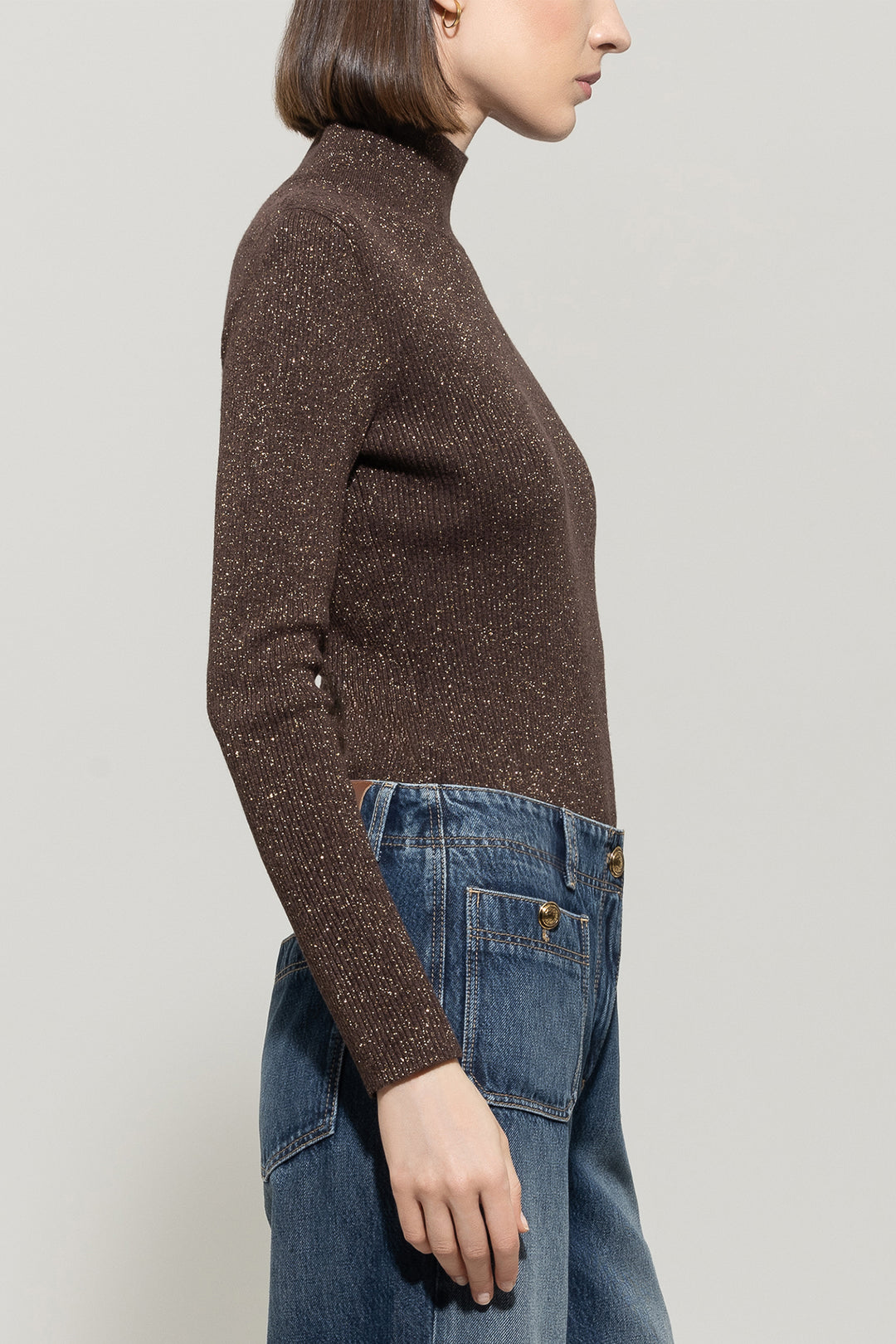 Person wearing a brown turtleneck sweater and blue jeans against a light gray background