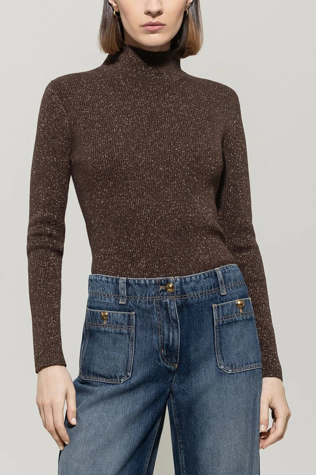 Person wearing a brown turtleneck sweater and blue jeans on a gray background