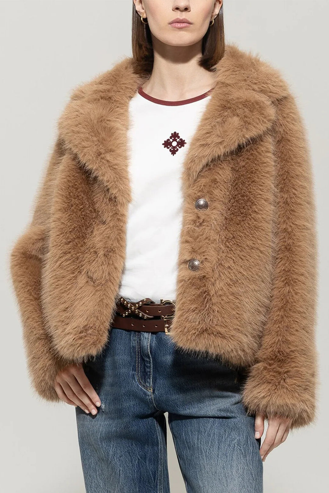 Person wearing a brown fluffy jacket, white shirt with red design, and blue jeans on a plain background