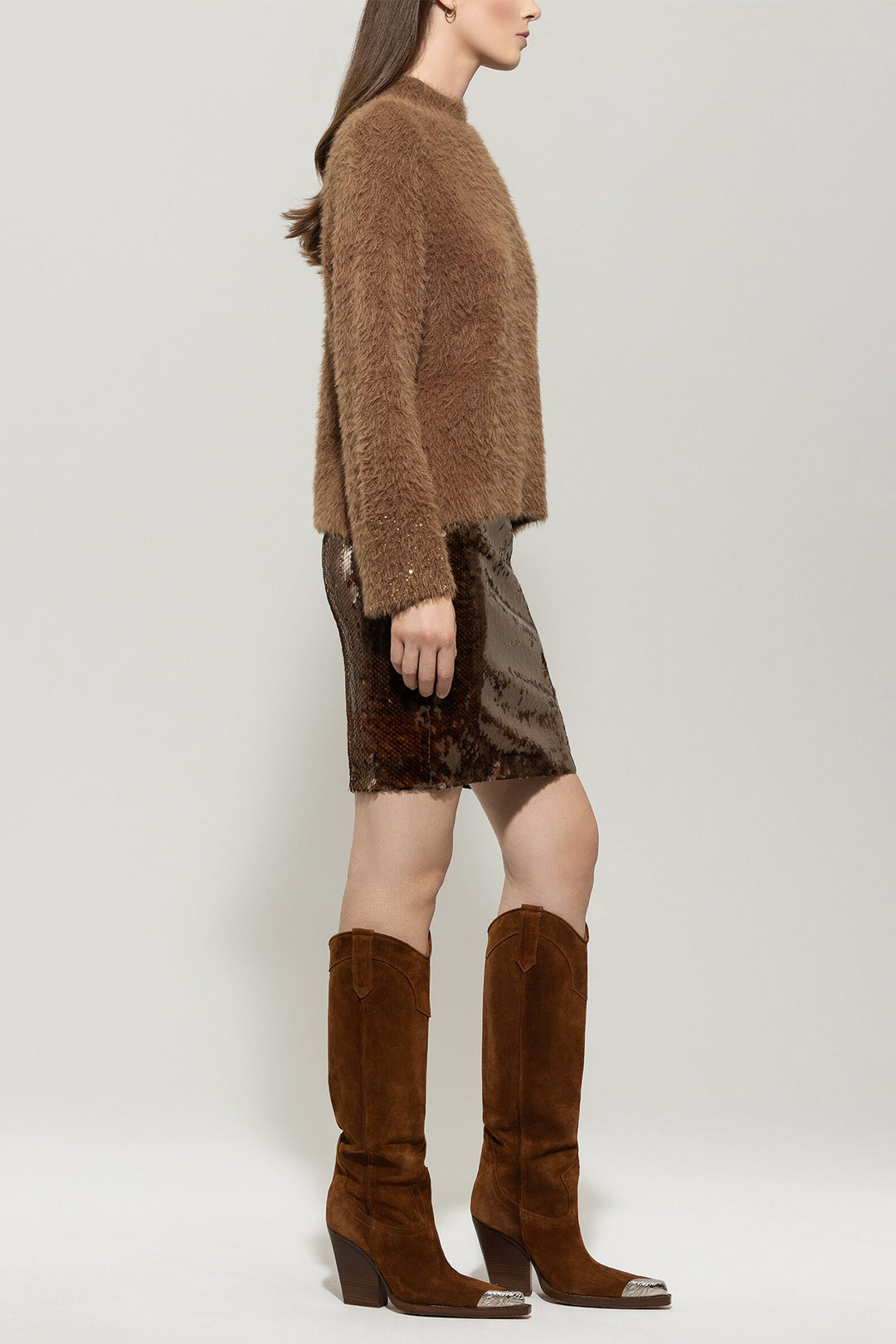 Woman wearing a brown fuzzy sweater, patterned skirt, and brown knee-high boots on a light gray background