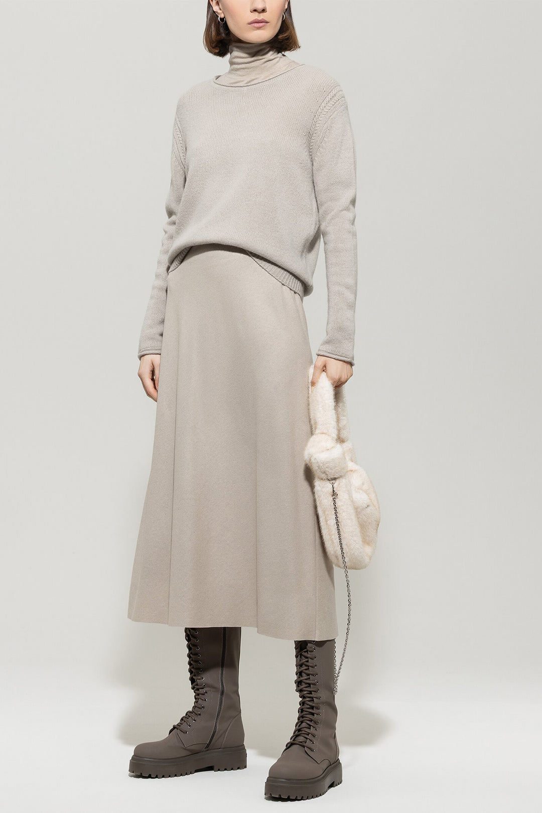 Person wearing a beige sweater, skirt, and boots holding a white bag on a light gray background