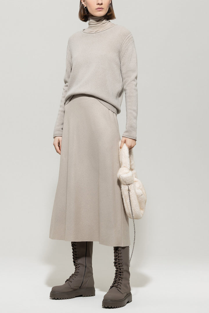 Person wearing a beige sweater, skirt, and boots holding a white bag on a light gray background