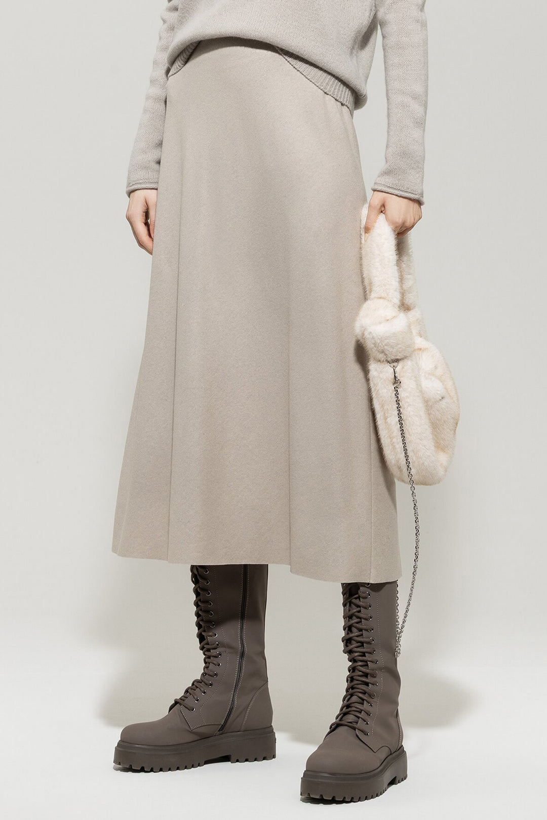 Person wearing a beige skirt, gray sweater, and brown boots holding a cream-colored bag against a white background