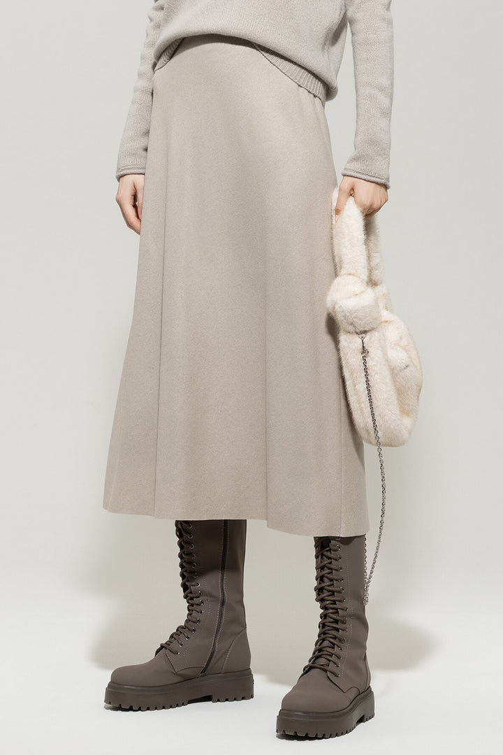 Person wearing a beige skirt, gray sweater, and brown boots holding a cream-colored bag against a white background