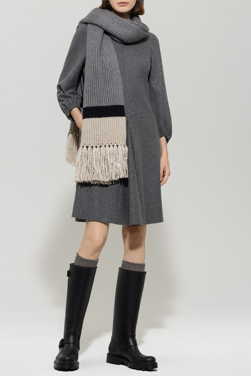 Person wearing a gray dress, black boots, and a long gray scarf with fringe on a white background