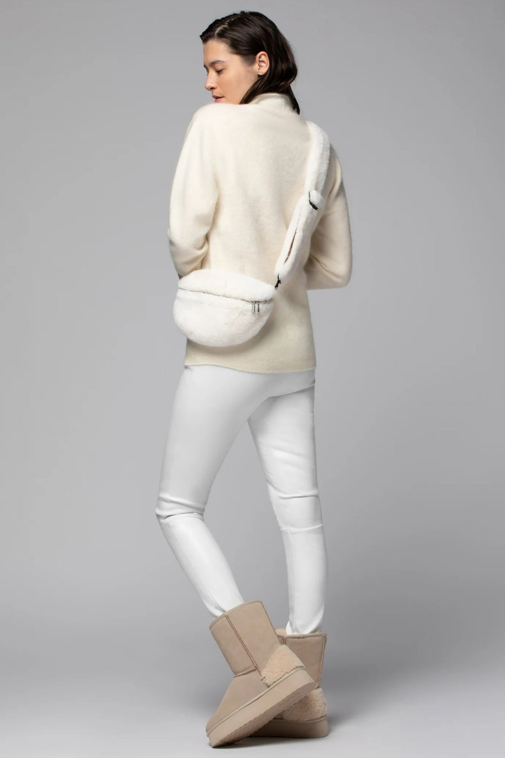 Woman wearing a cream sweater, white pants, and beige boots on a gray background