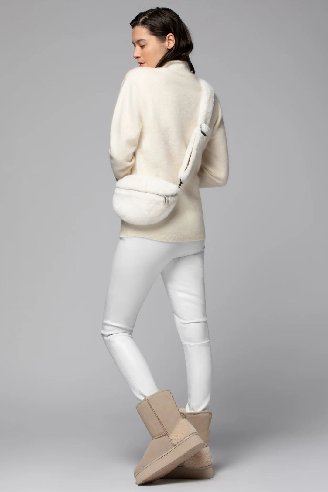 Woman wearing a cream sweater, white pants, and beige boots on a gray background