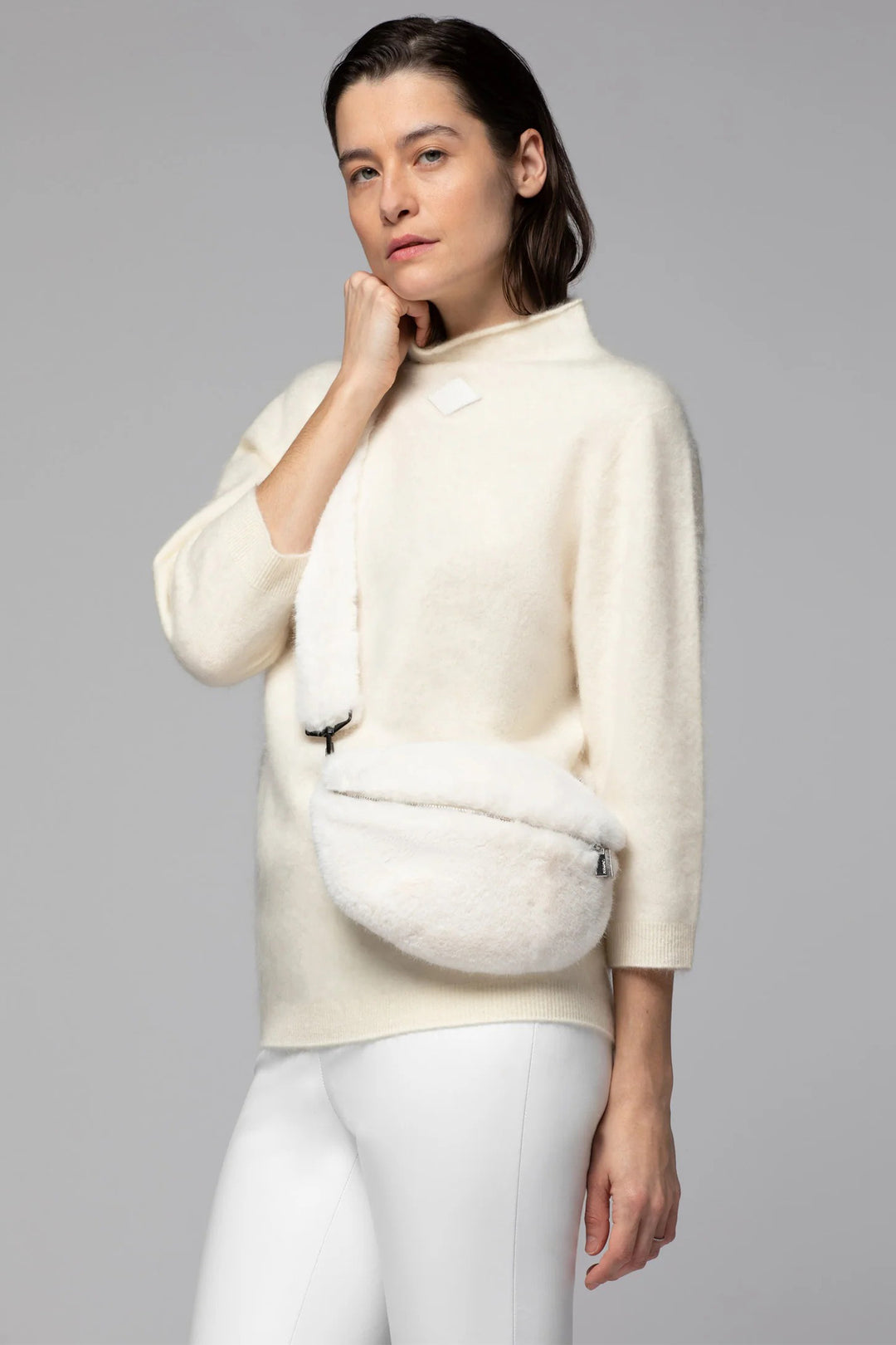 Woman wearing a cream-colored sweater with a fur-lined pocket against a gray background