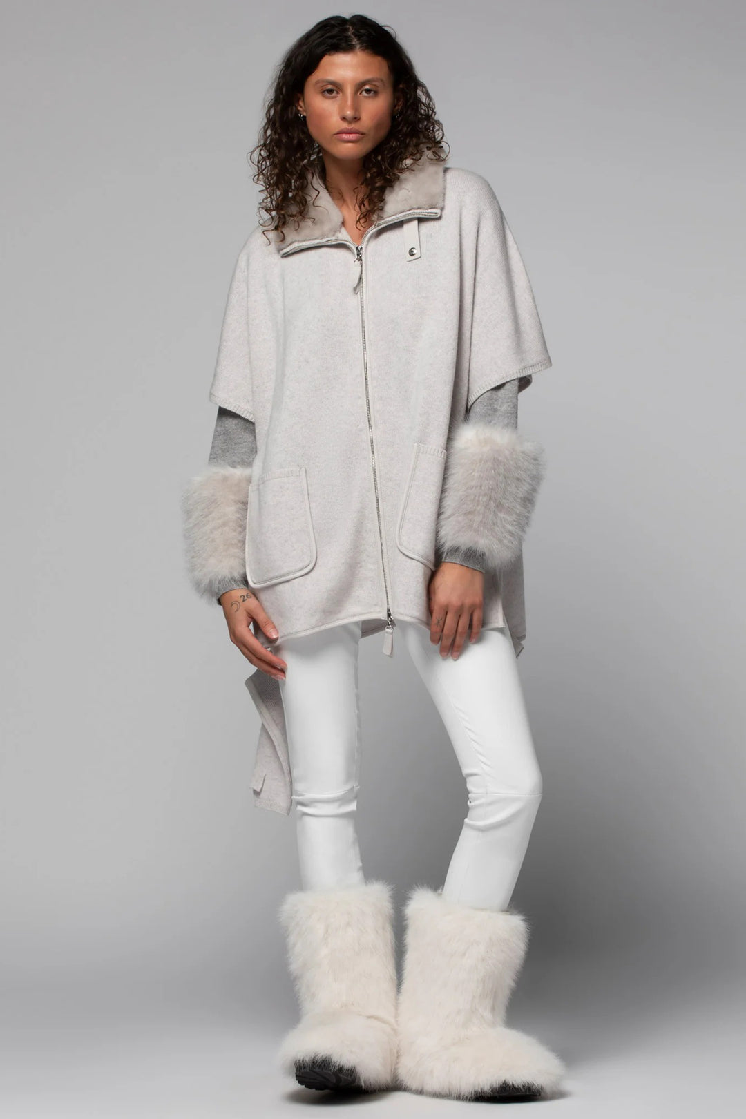 Woman wearing a light gray coat with fur trim, white pants, and white boots on a gray background