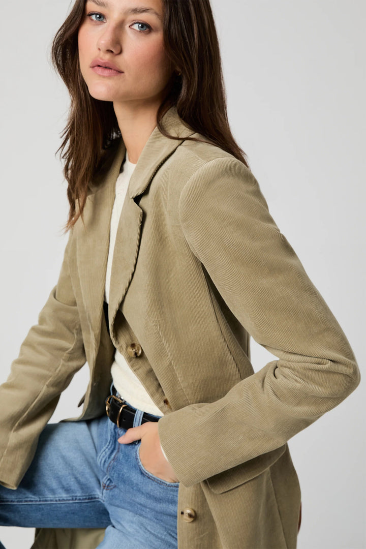 Woman wearing a beige blazer over a white shirt with blue jeans on a plain background