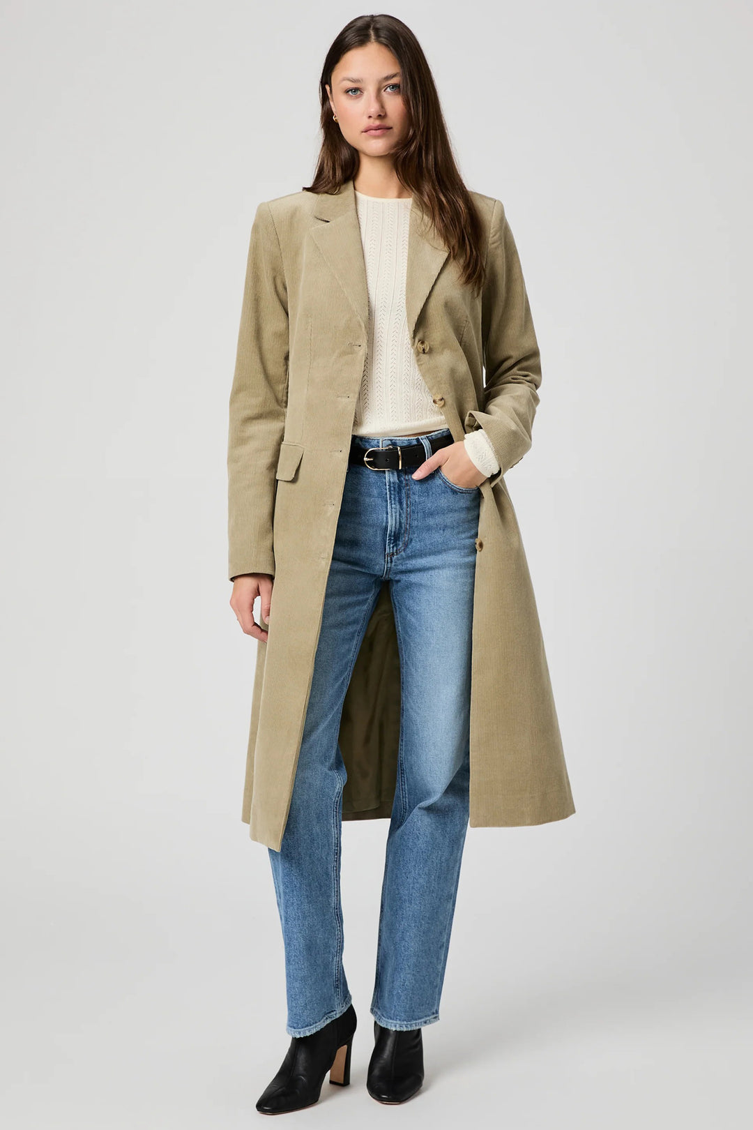 Woman wearing a beige coat, white shirt, blue jeans, and black shoes on a white background