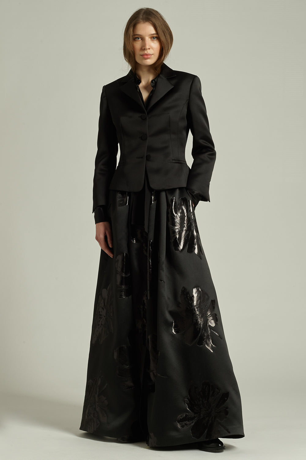 Person wearing a black suit with a long black dress underneath on a plain background