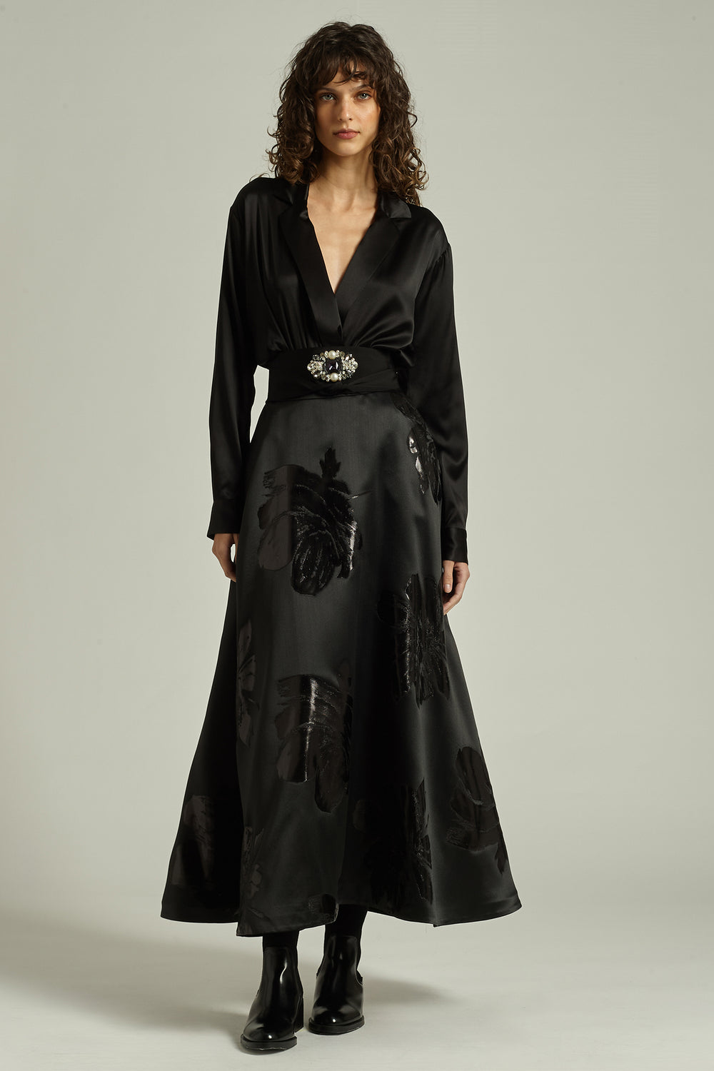 Woman wearing a black outfit with a belt and long skirt against a neutral background
