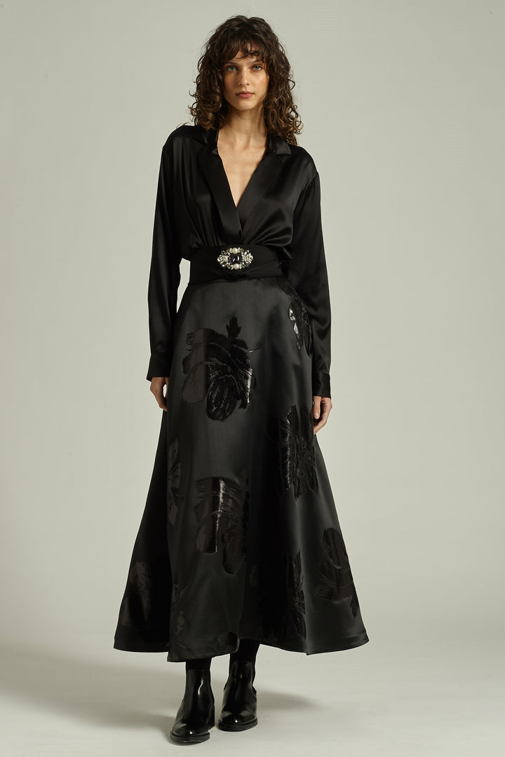 Woman wearing a black outfit with a belt and long skirt against a neutral background