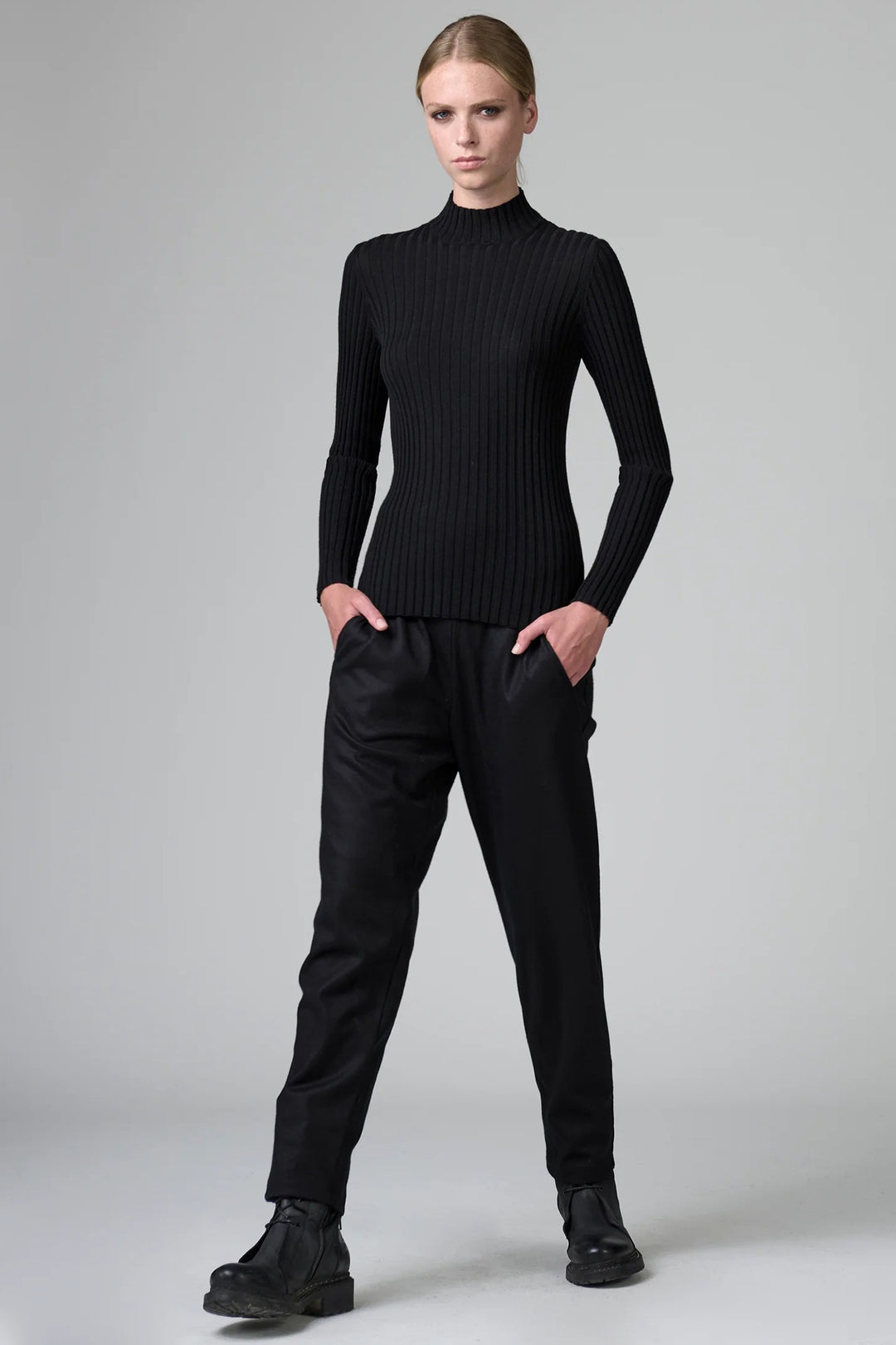 Person wearing a black ribbed turtleneck sweater and black pants on a gray background