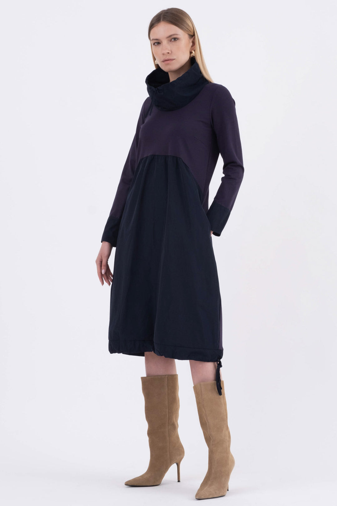 Woman wearing a long dark dress with a cowl neck and brown boots on a white background